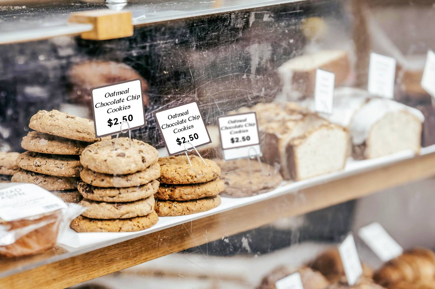 Ein Regal mit Keksen und Brotscheiben hinter Glas. Schilder zeigen Preise: 'Oatmeal Chocolate Chip Cookies $2,50' und 'Oatmeal Cookies $2,50'.