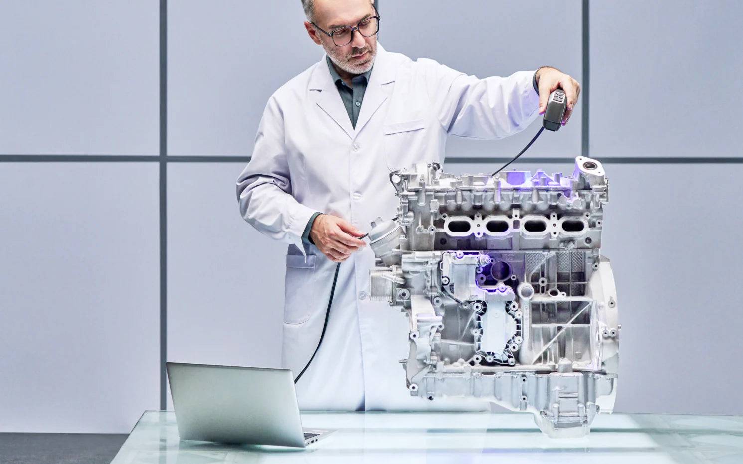 Ein Wissenschaftler in weißem Kittel untersucht einen transparenten Motorblock auf einem Tisch mit einem Laptop daneben.