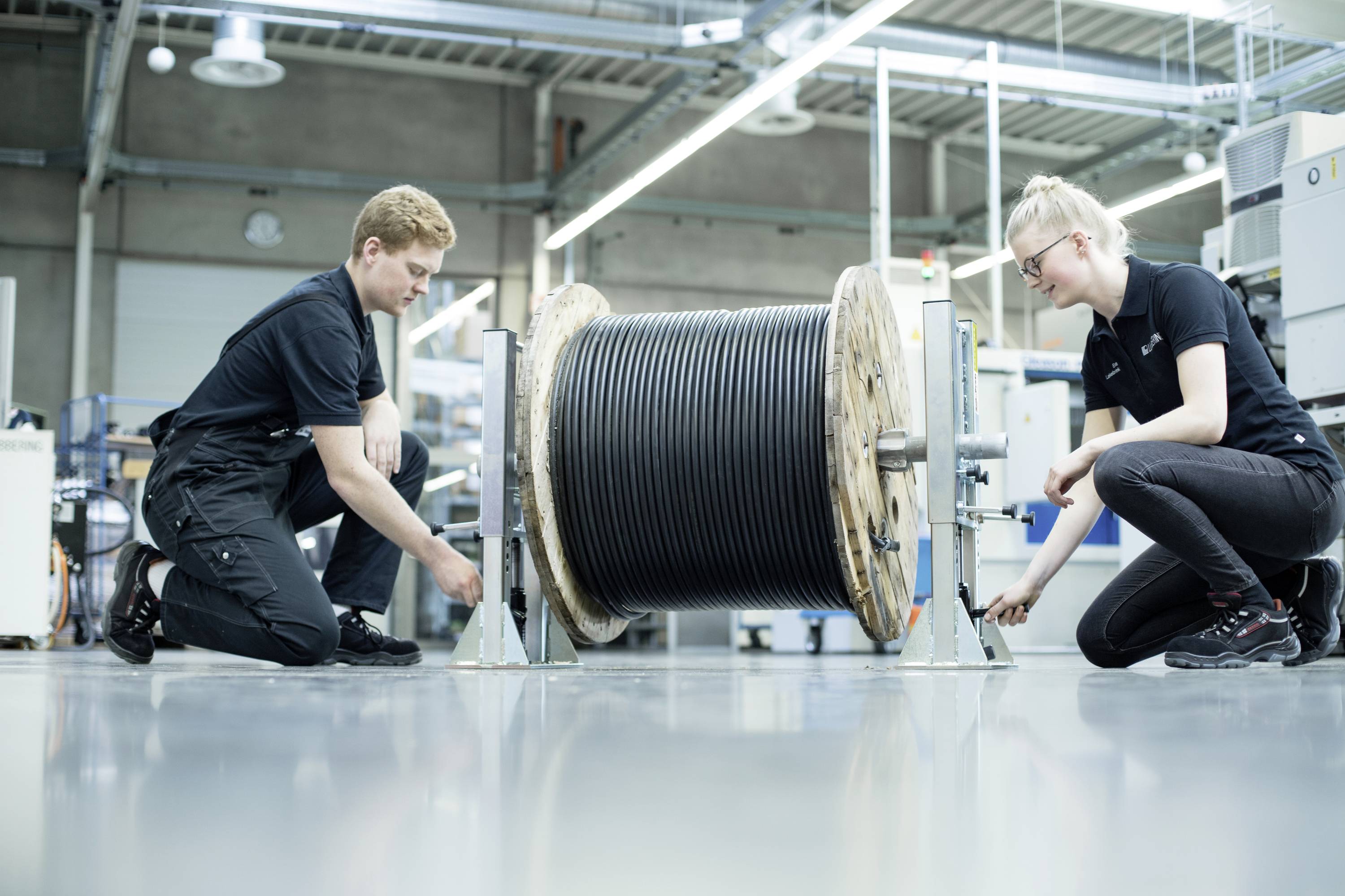 Zwei Personen in Arbeitskleidung knien auf einem Industrieboden und überprüfen eine große Kabeltrommel in einer Werkshalle.