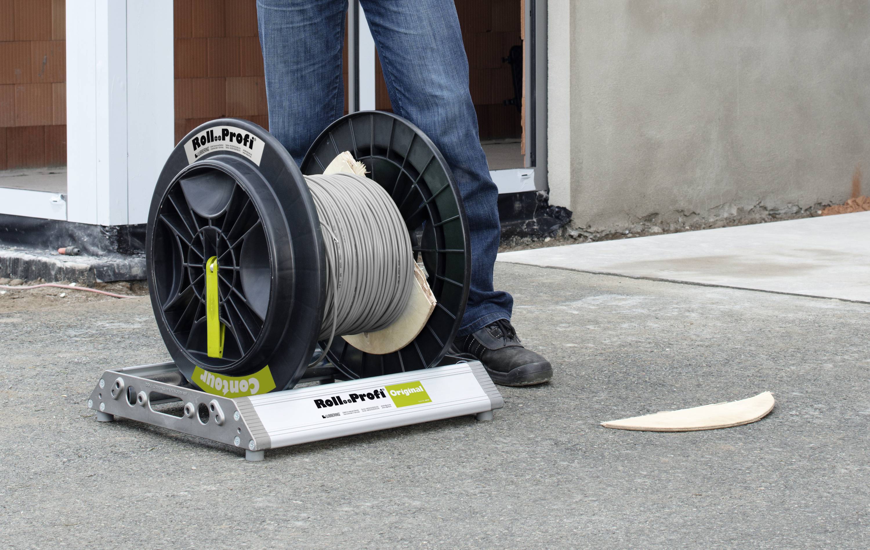 Eine Kabeltrommel namens 'Roll-Profi Original' steht auf einer metallischen Halterung auf einem Betonboden. Eine Person im Hintergrund.