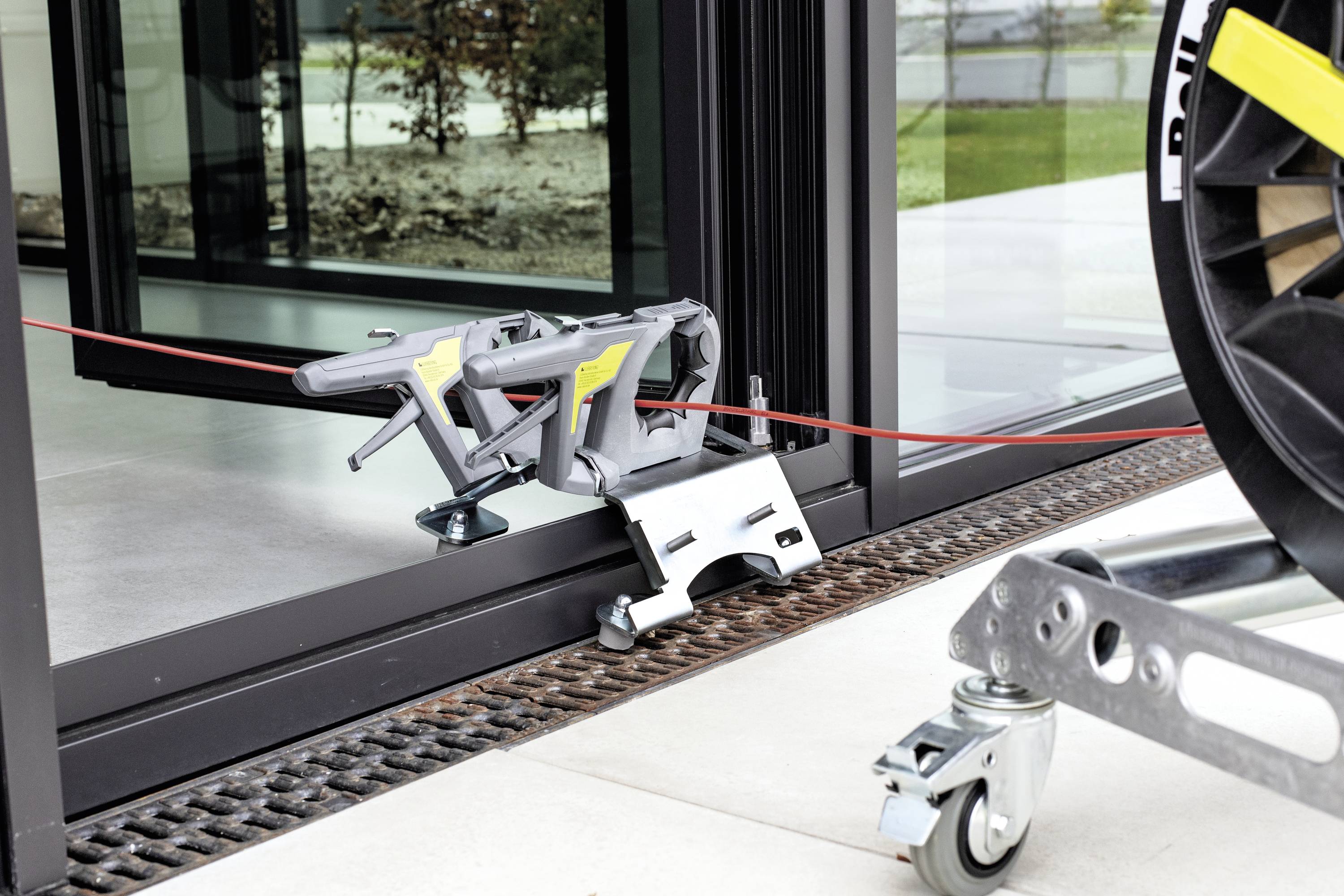 Ein Gerät zieht ein rotes Kabel durch einen offenen Schiebetürrahmen. Im Vordergrund ein Wagenrad. Die Szene zeigt ein Kabelverlegeprozess.