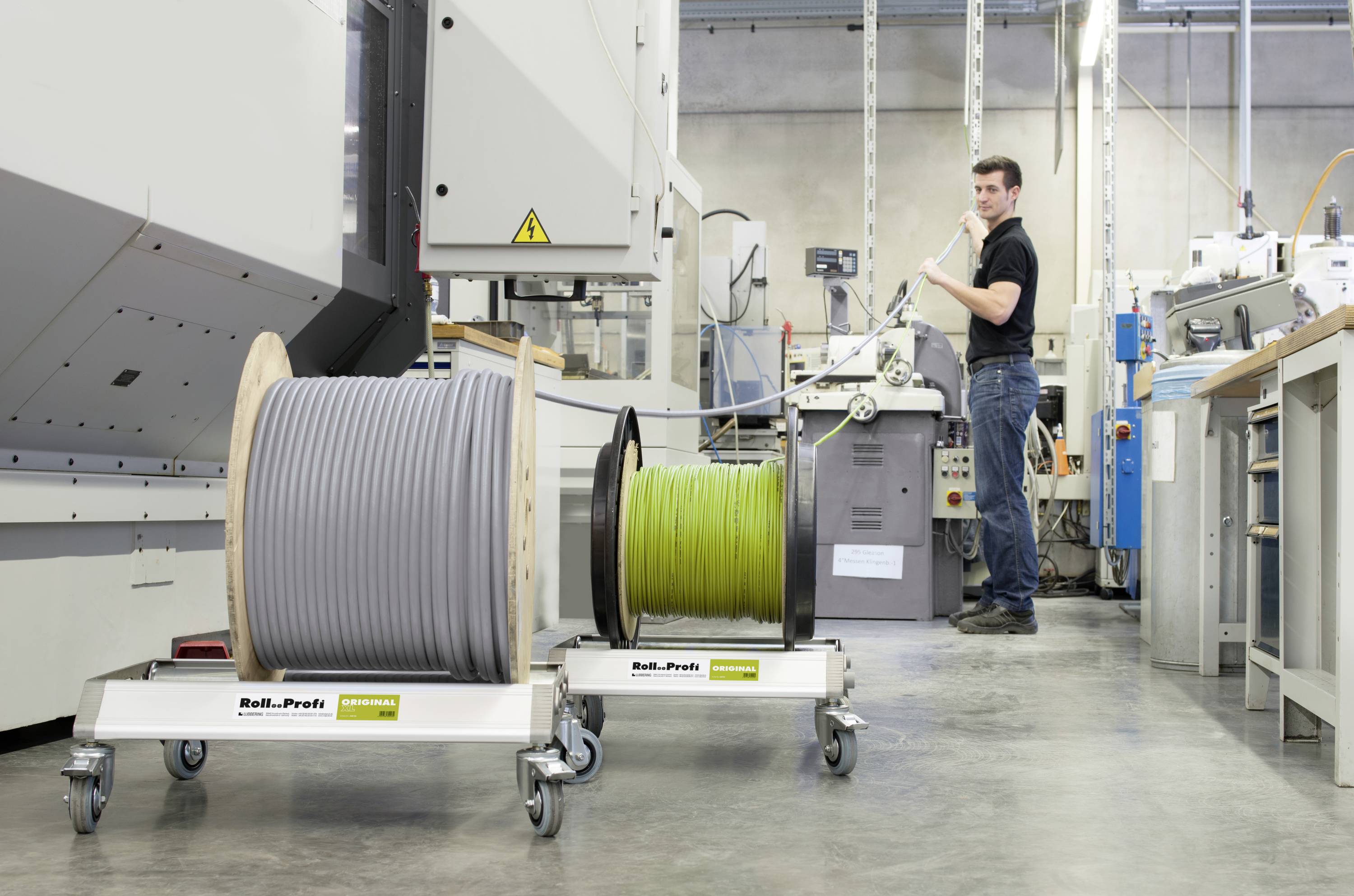 Ein Mann arbeitet in einer Fabrikhalle mit zwei großen Kabeltrommeln auf Wagen. Im Hintergrund sind Maschinen und Arbeitsgeräte zu sehen.