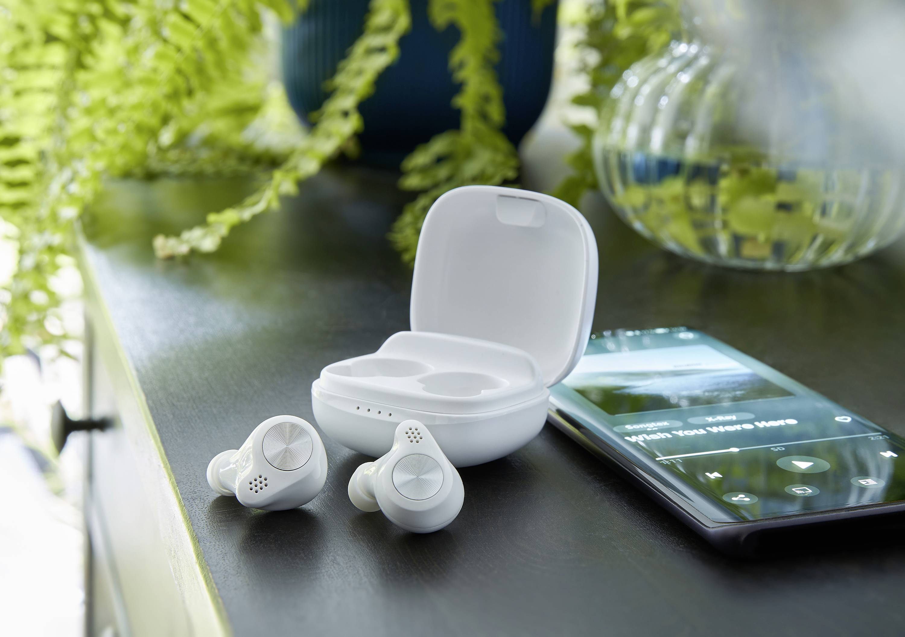 Kabellose weiße Ohrhörer in einer Ladehülle mit einem Smartphone, das Musik abspielt, auf einem Tisch neben grünen Pflanzen.