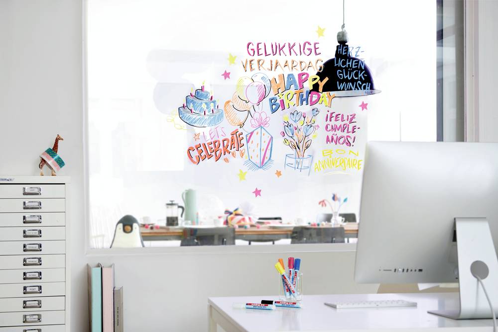 Ein Büro mit einem Schreibtisch, auf dem Stifte und ein Computer stehen. An der Glasscheibe kleben bunte Geburtstagswünsche.