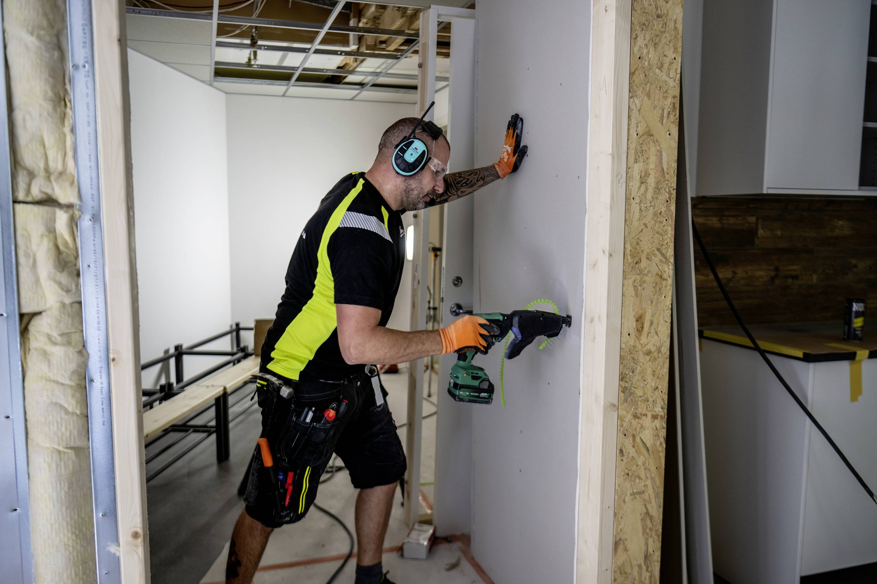 Ein Handwerker in Sicherheitskleidung und Gehörschutz schraubt eine Platte an eine Holzwand. Im Hintergrund sieht man Möbel und Werkzeuge.