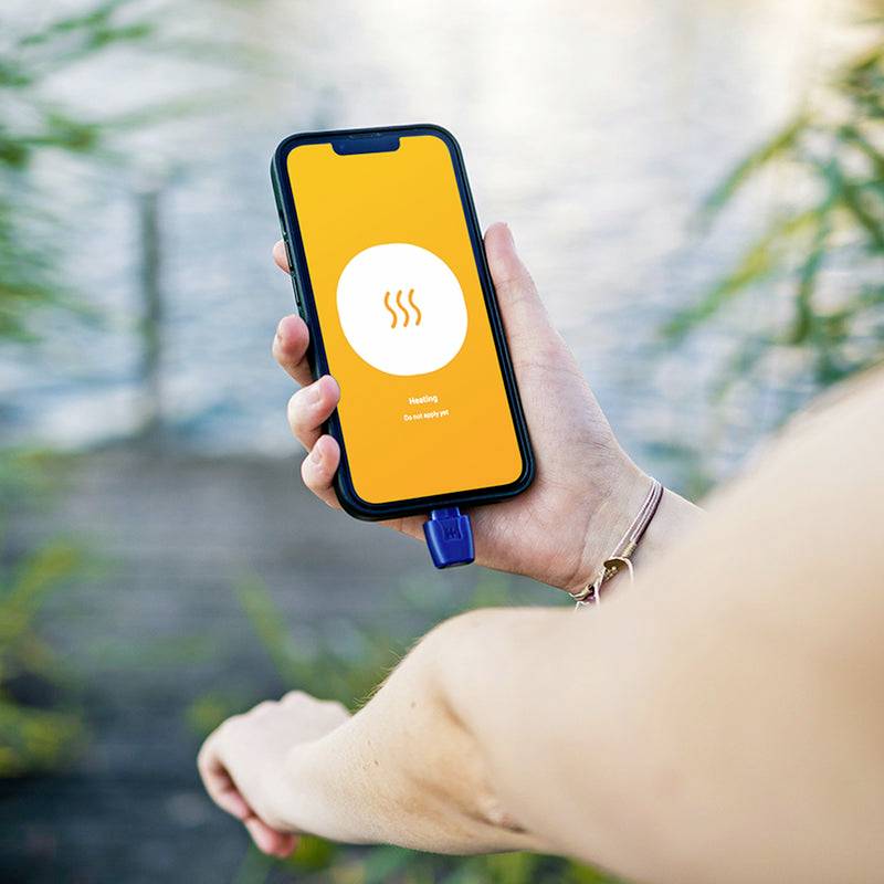 Eine Person hält ein Smartphone mit einer App mit der Aufschrift 'heating' auf dem Bildschirm. Im Hintergrund ist Wasser zu sehen.
