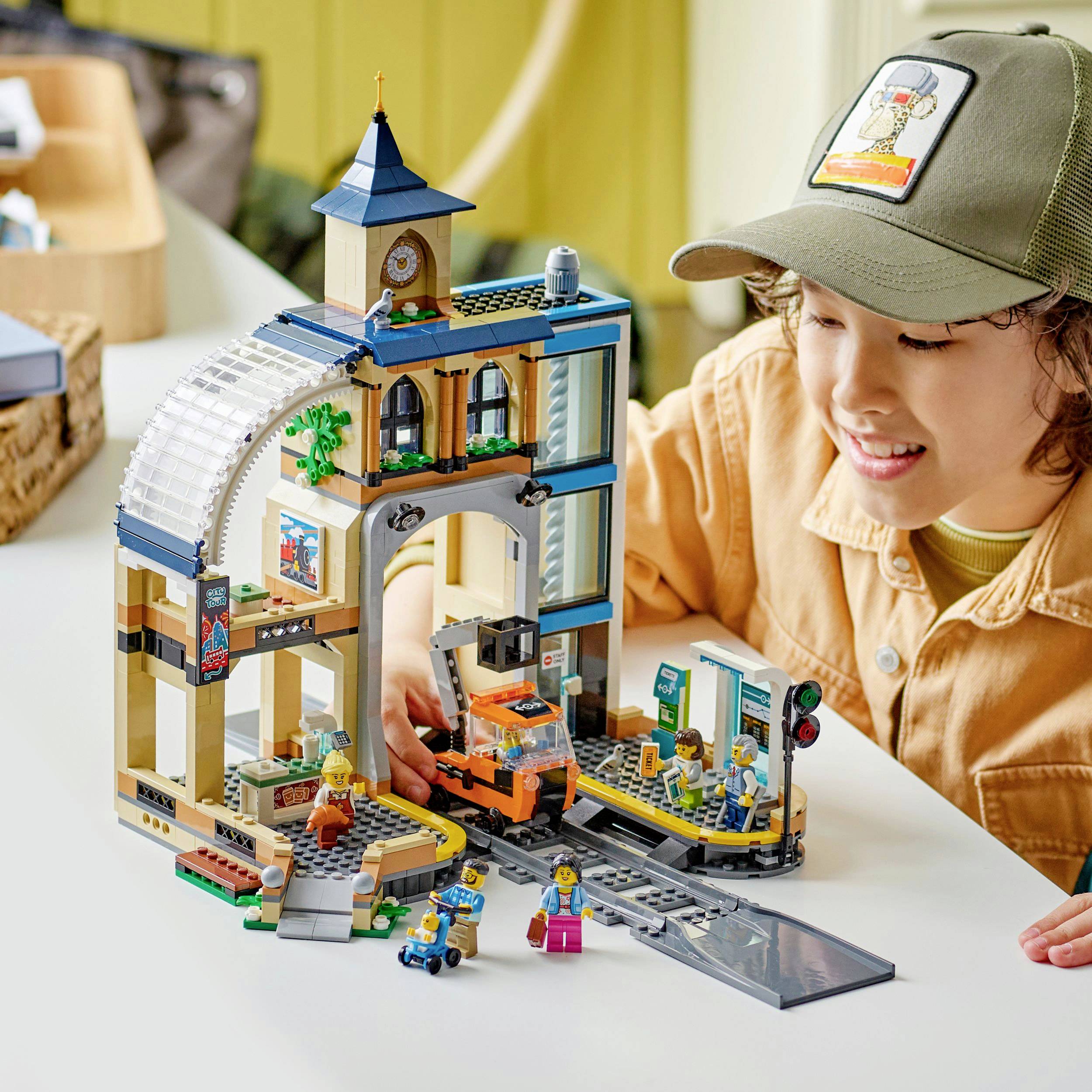 Ein Kind spielt mit einer Lego-Bausatz, der einen Bahnhof darstellt, mit Zug, Figuren, und Bahnstrecke. Es wirkt konzentriert und glücklich.