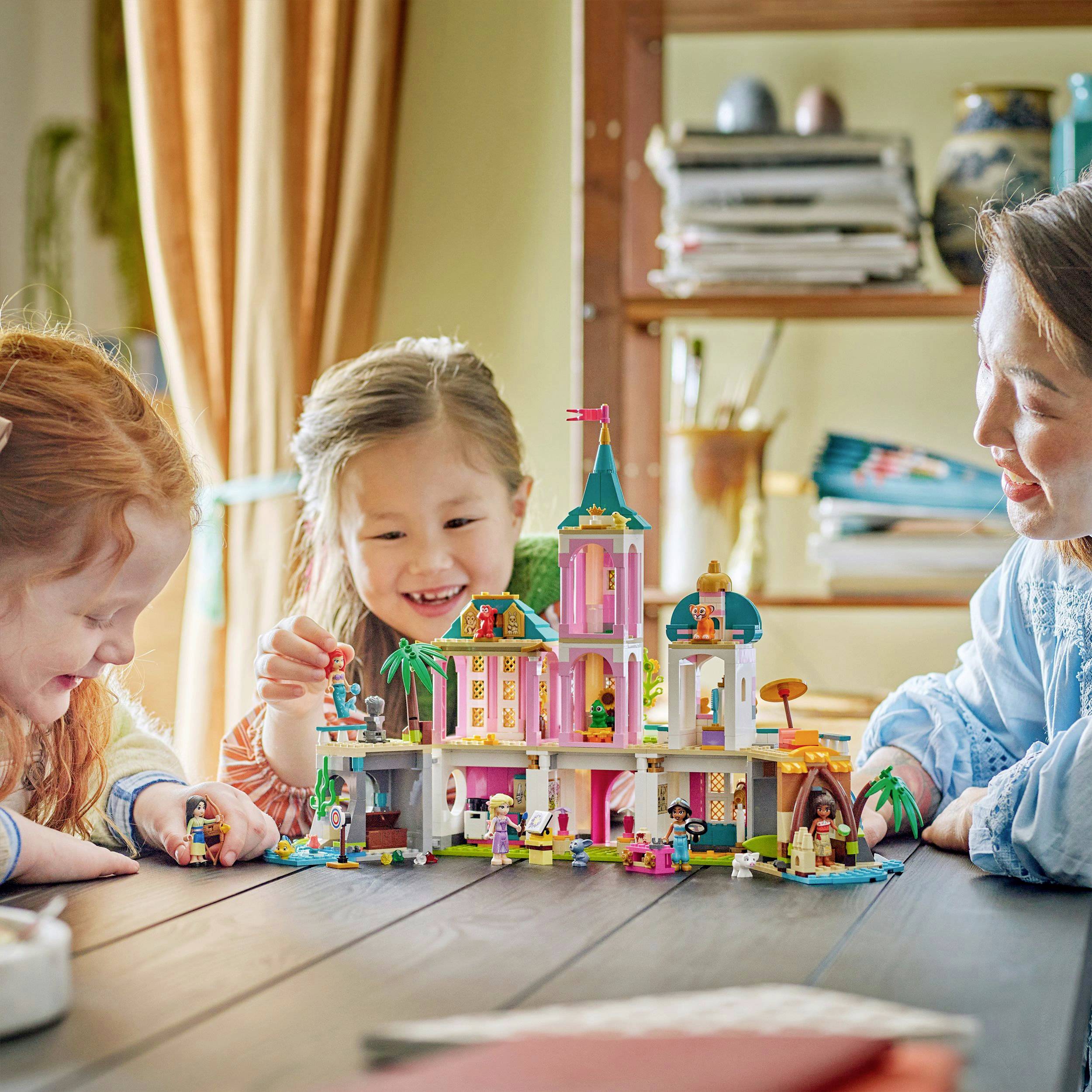 Drei lachende Kinder und eine Frau spielen mit einem bunten, fantasievollen Spielzeugschloss auf einem Tisch in einem gemütlichen Raum.
