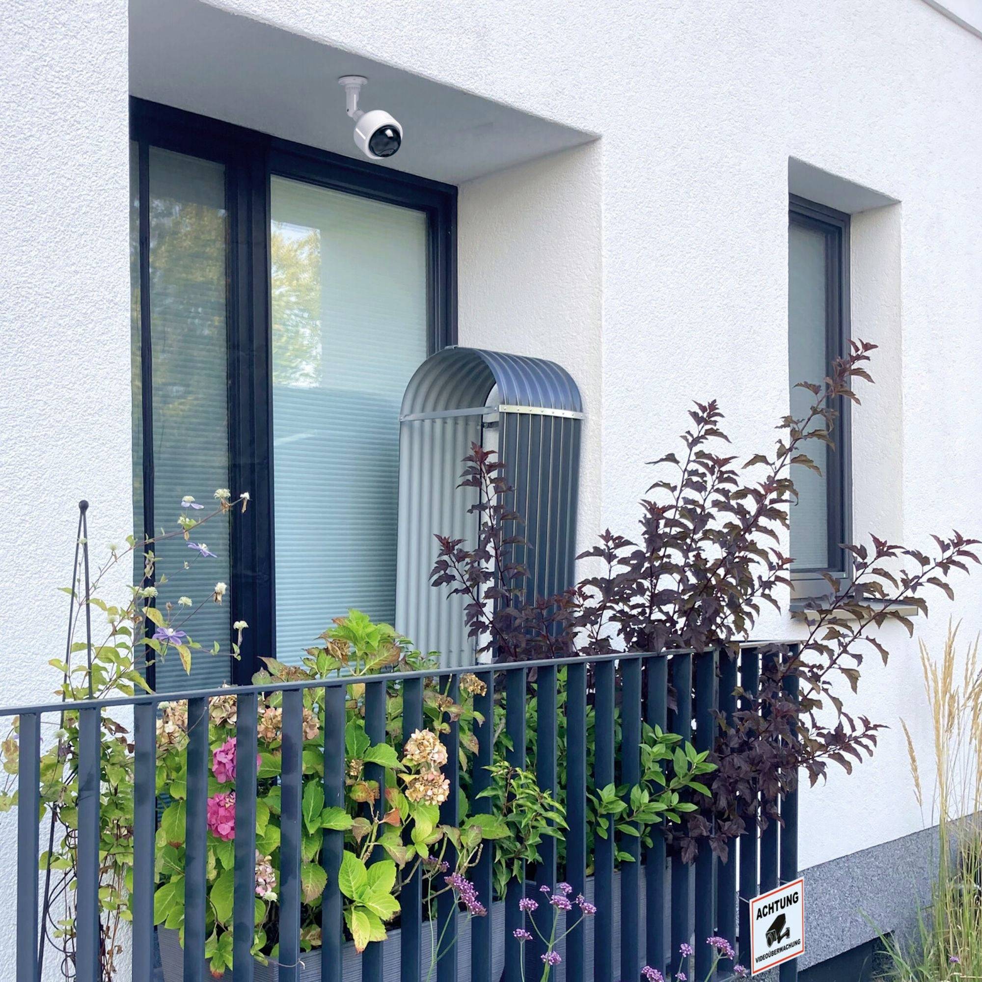 Überwachungsgerät an einem modernen Gebäude, oberhalb eines Balkons mit Pflanzen; Schild 'Achtung' weist auf Videoüberwachung hin.