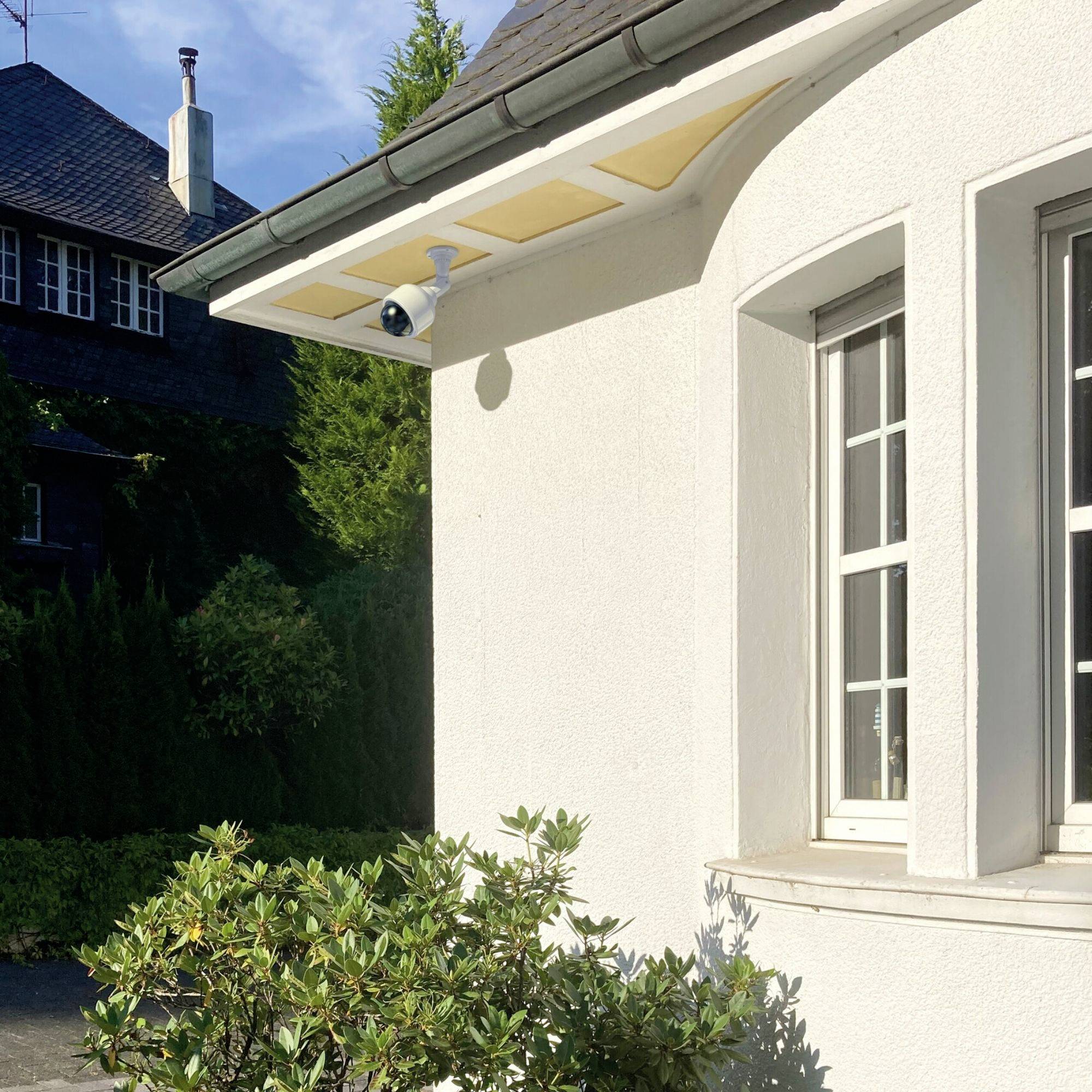 Ein weißes Haus mit einem Fenster und Überwachungskamera unter dem Dachvorsprung. Dichte Büsche und Bäume im Hintergrund.