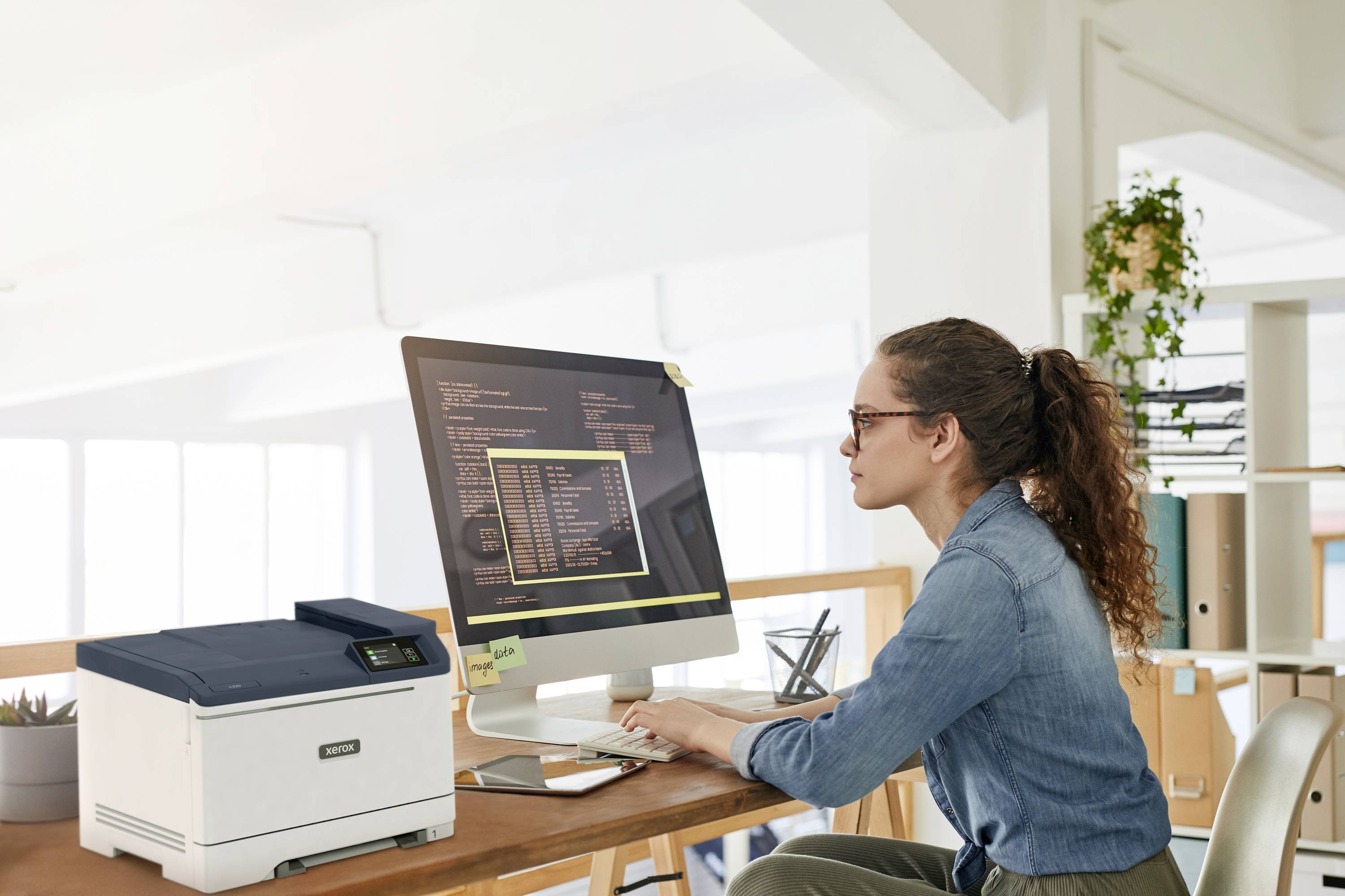 Eine Person sitzt an einem Schreibtisch und arbeitet konzentriert an einem Computer mit Code auf dem Bildschirm. Daneben steht ein Drucker.