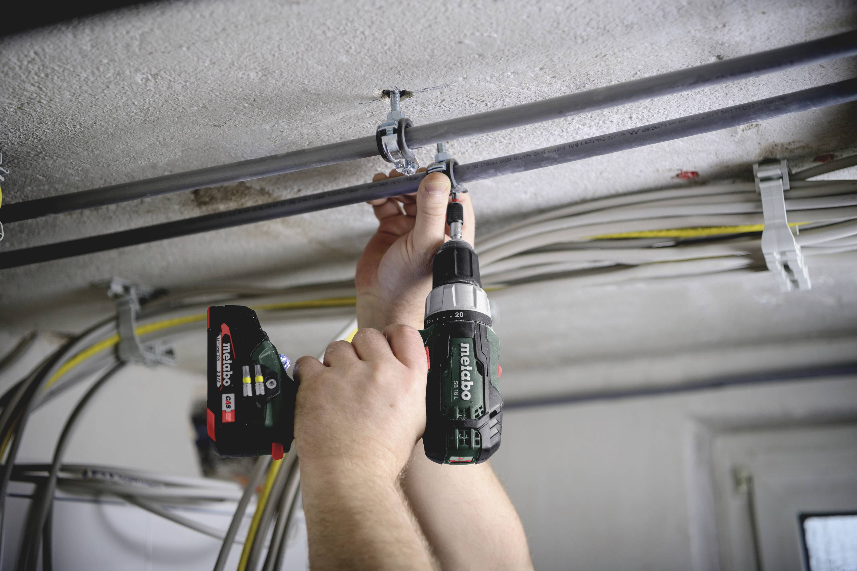 Person montiert Rohre an der Decke mit einem Akkuschrauber. Zahlreiche Kabel sind ebenfalls an der Decke befestigt.