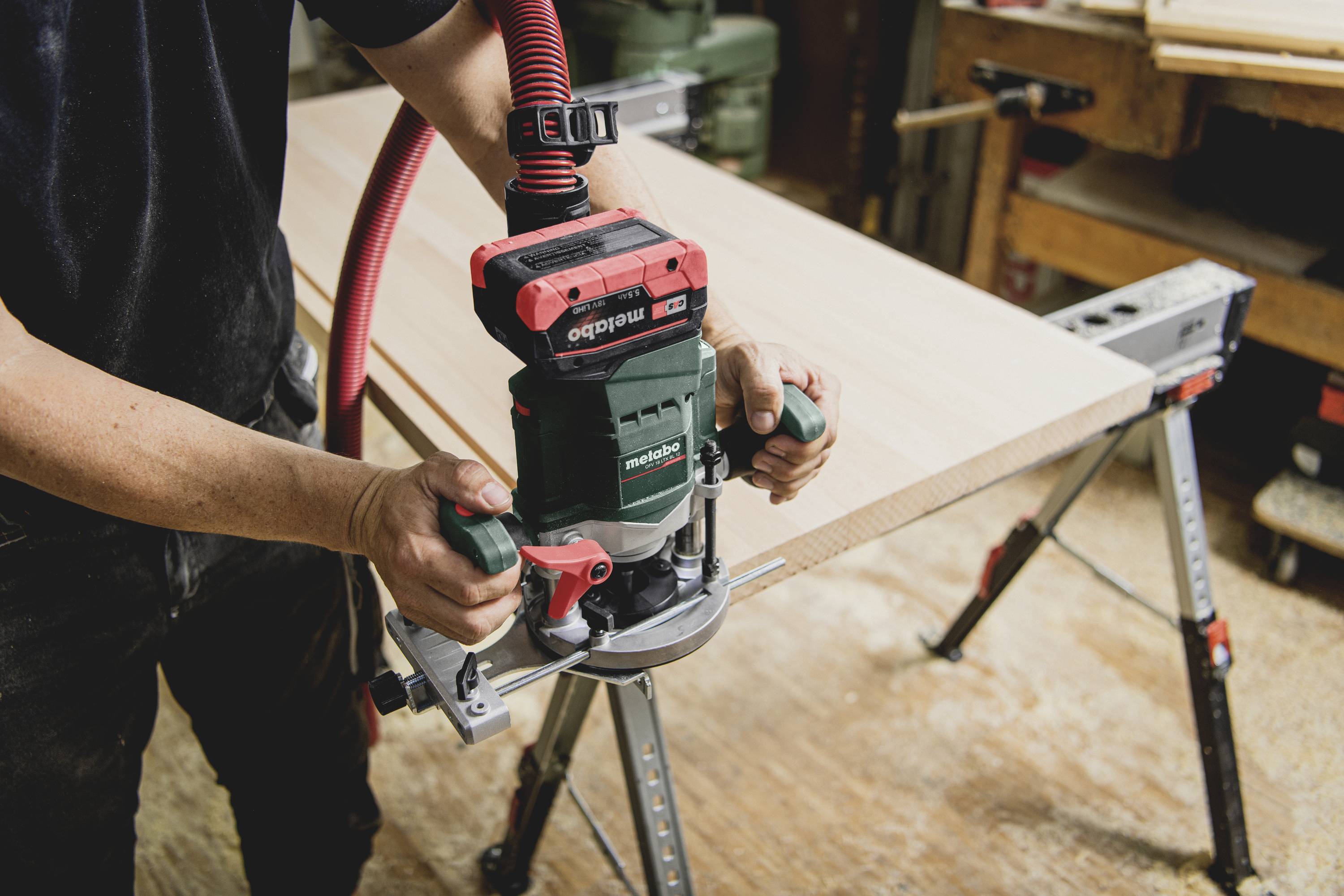 Eine Person fräst mit einer Oberfräse eine Holzplatte in einer Werkstatt, wobei sie das Gerät mit beiden Händen führt.