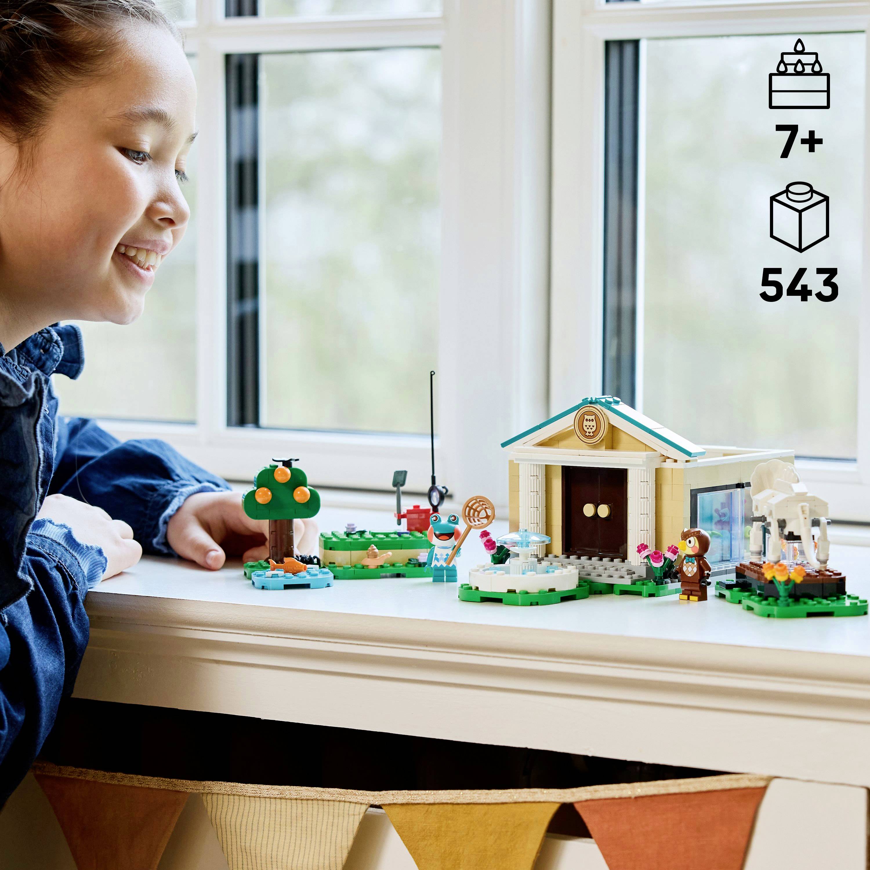 Ein Kind spielt mit einem LEGO-Set auf einem Tisch neben einem Fenster, wobei ein Haus, ein Baum und verschiedene kleine Figuren zu sehen sind. Geeignet für Kinder ab 7 Jahren.