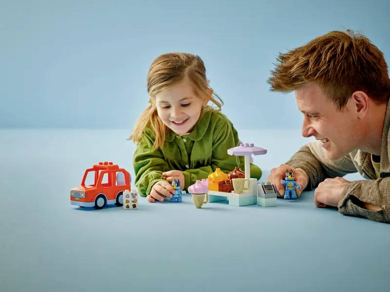 Ein Kind und ein Erwachsener spielen mit bunten Spielfiguren und einem kleinen Spielzeugfahrzeug auf einer hellblauen Oberfläche, lächeln und sind gemeinsam beschäftigt.