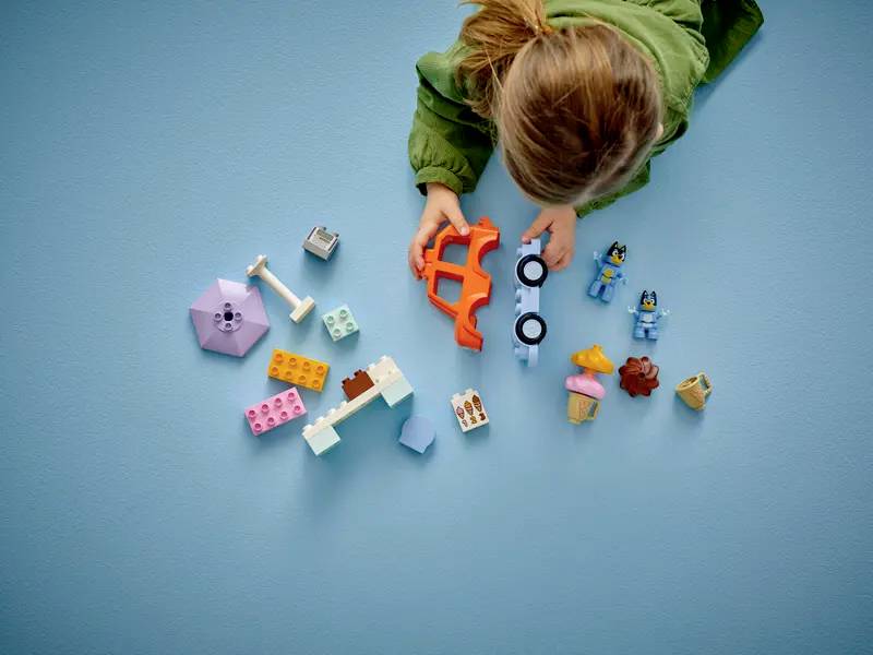 Ein Kind in einem grünen Pullover spielt mit bunten Spielblöcken und Figuren auf einem blauen Boden und konzentriert sich darauf, ein kleines Fahrzeug zusammenzubauen.