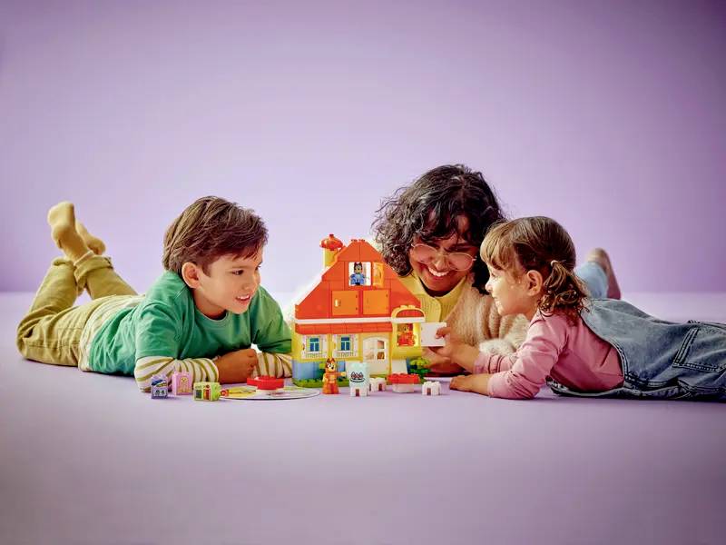 Ein fröhlicher Erwachsener und zwei Kinder spielen mit einem Spielhaus auf einem hellvioletten Hintergrund und zeigen dabei kreatives Spiel und gemeinsame Bindungszeit.