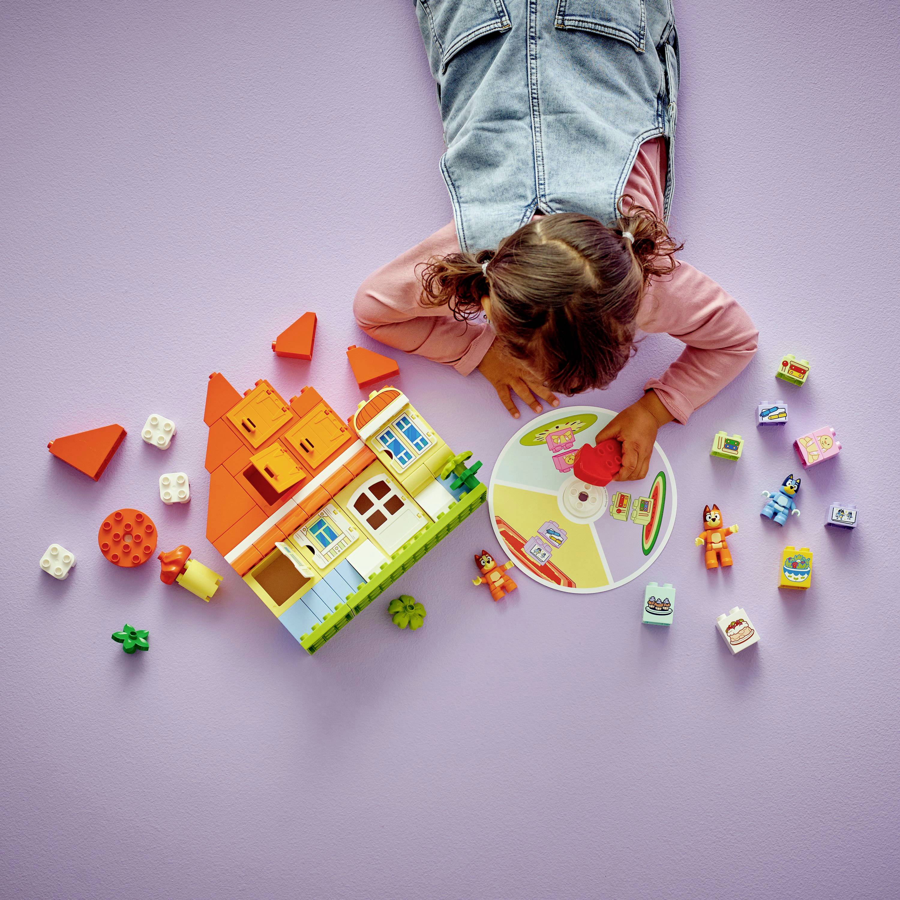 Ein Kind spielt auf einem lila Boden mit bunten Bauklötzen, einem Puppenhaus und kleinen Figuren und schafft dabei eine fantasievolle und spielerische Szene.