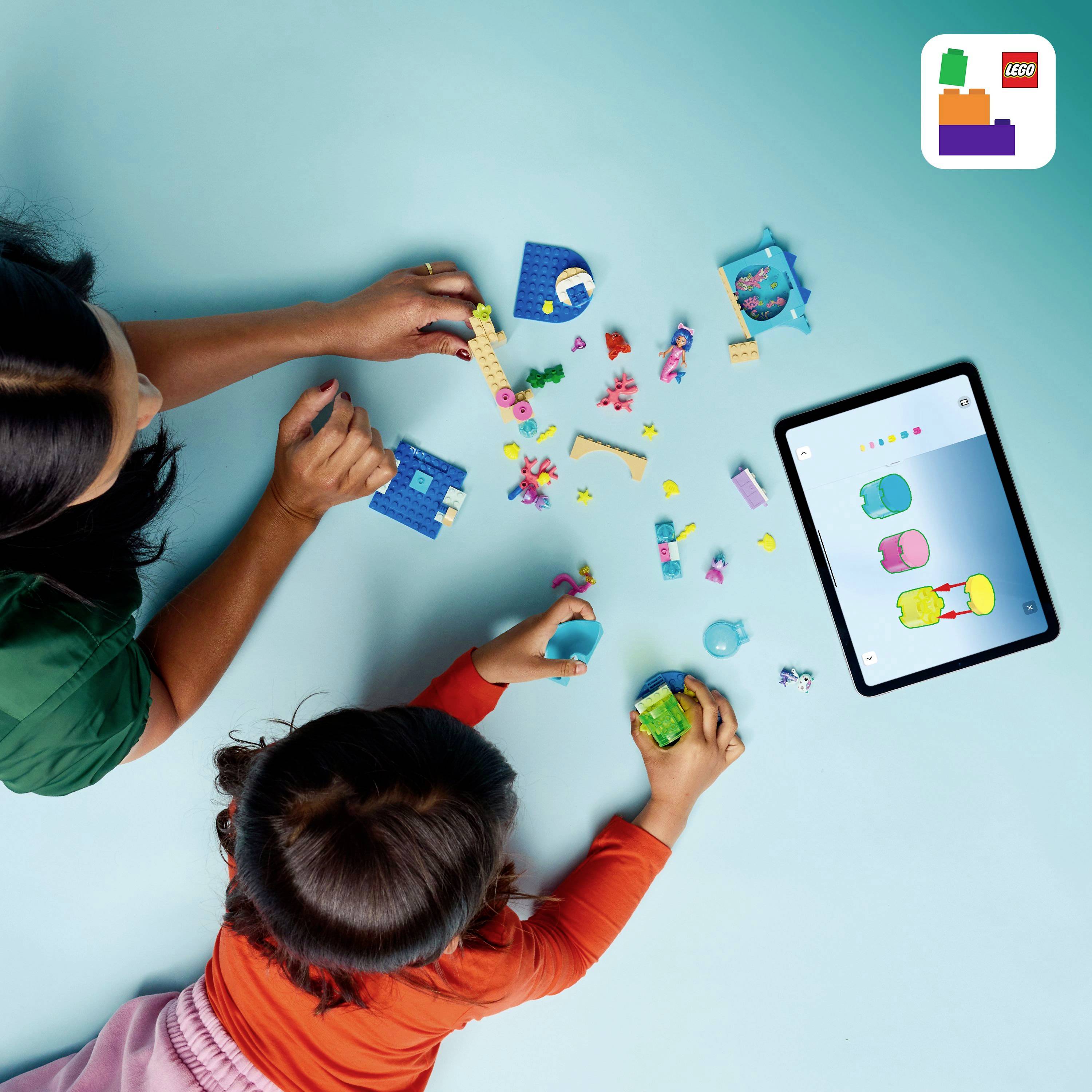 Ein Kind und ein Erwachsener spielen mit bunten LEGO-Steinen auf einem Tisch. Ein iPad zeigt eine zugehörige LEGO-App.