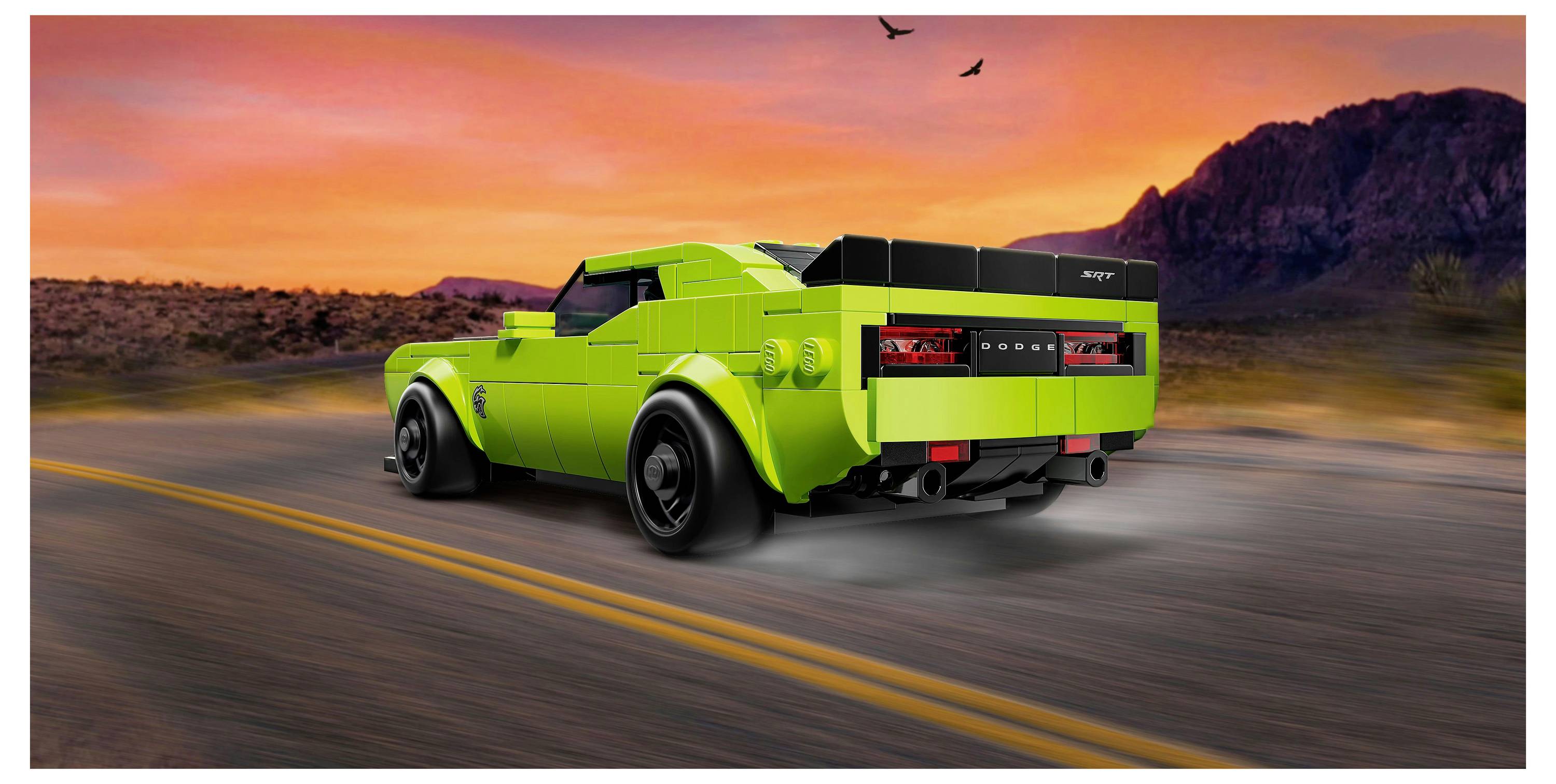 Ein lebendiges grünes Spielzeugauto auf einer Straße, vor einem dramatischen Sonnenuntergang mit Bergen im Hintergrund, weckt Assoziationen von Geschwindigkeit und Abenteuer.