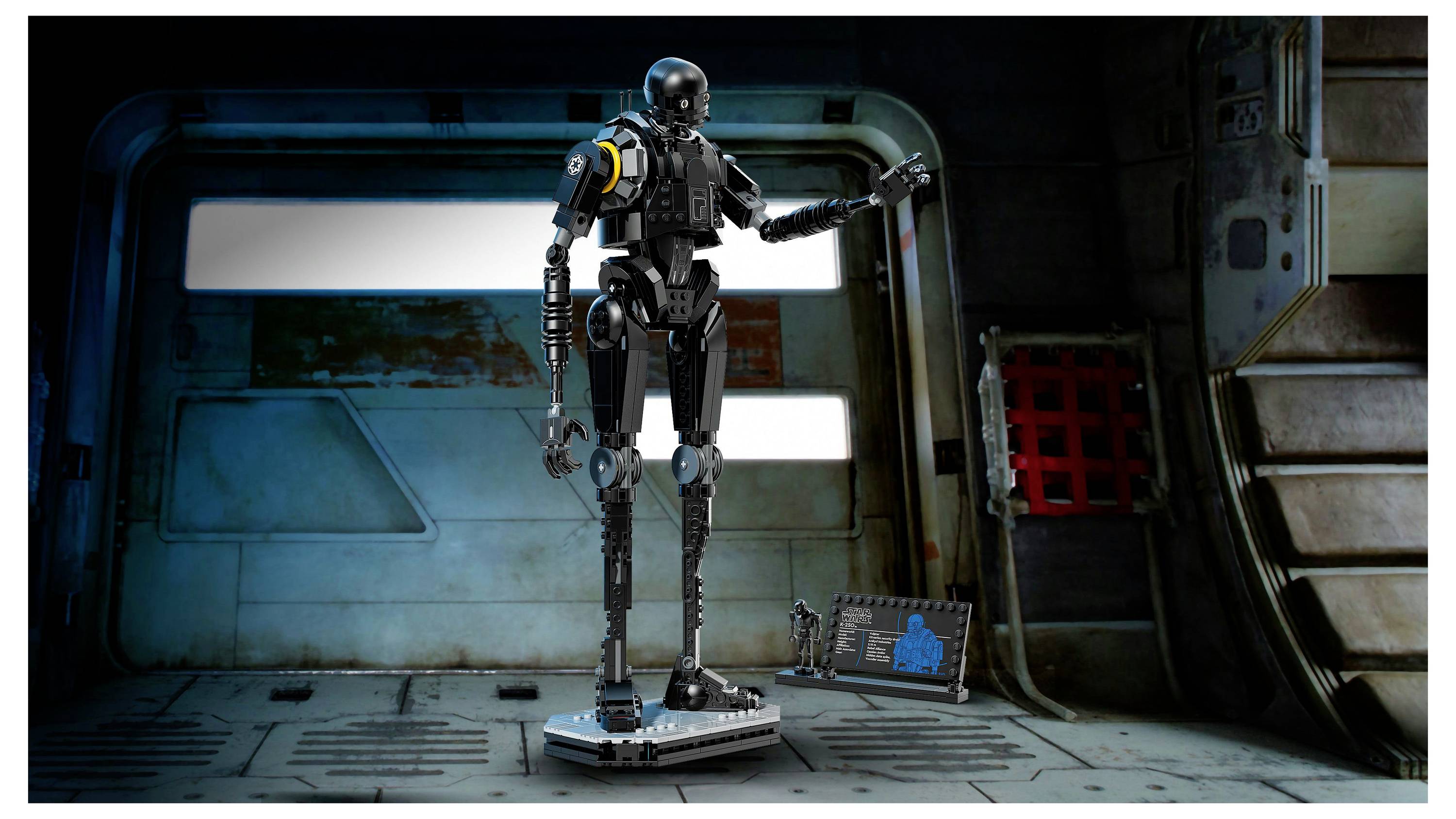 Ein großer, schwarzer humanoider Roboter steht in einem düster beleuchteten Industrieraum und gestikuliert mit einem Arm. Ein kleiner Bildschirm zeigt ein blaues Schematisches Diagramm.