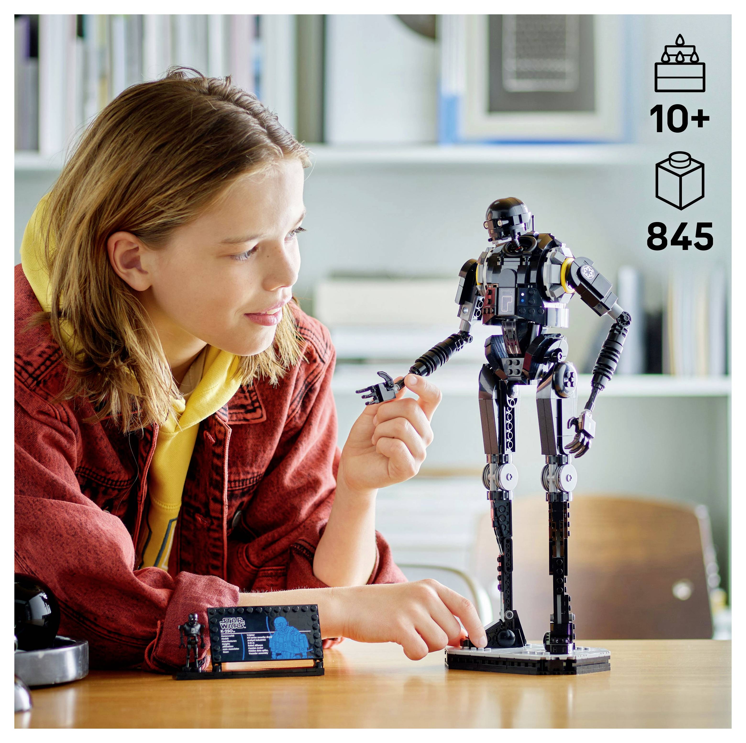 Ein junger Mensch montiert an einem Tisch ein Robotermodell mit Roboterteilen und ist konzentriert darauf bedacht, ein Teil zu positionieren. Im unscharfen Hintergrund befindet sich ein Bücherregal.