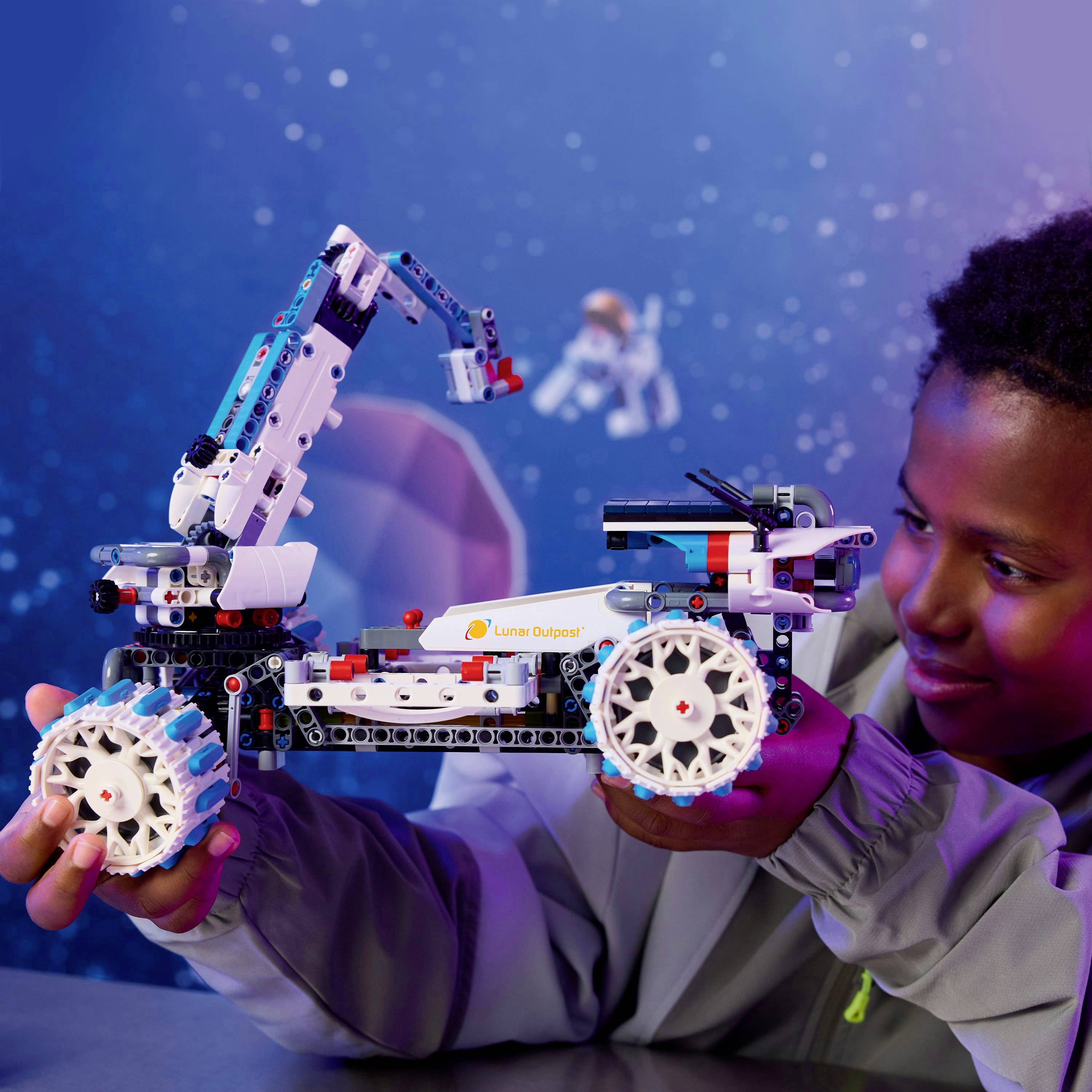 Ein Kind baut ein weiß-blaues Robotermodell mit großen Rädern und einem Arm, beschriftet mit 'Lunar Outpost'. Eine kleine Astronautenfigur schwebt im Hintergrund.