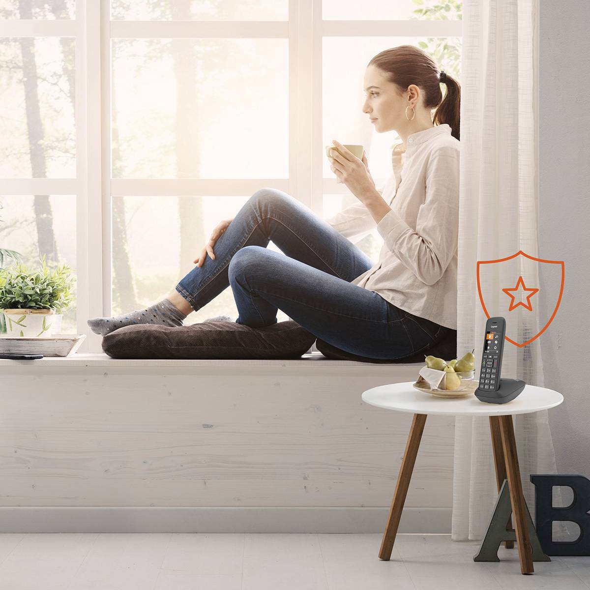 Eine Frau sitzt auf einer Fensterbank und trinkt Kaffee. Sonnenlicht fällt durch das Fenster. Neben ihr steht ein Tisch mit einem Telefon.