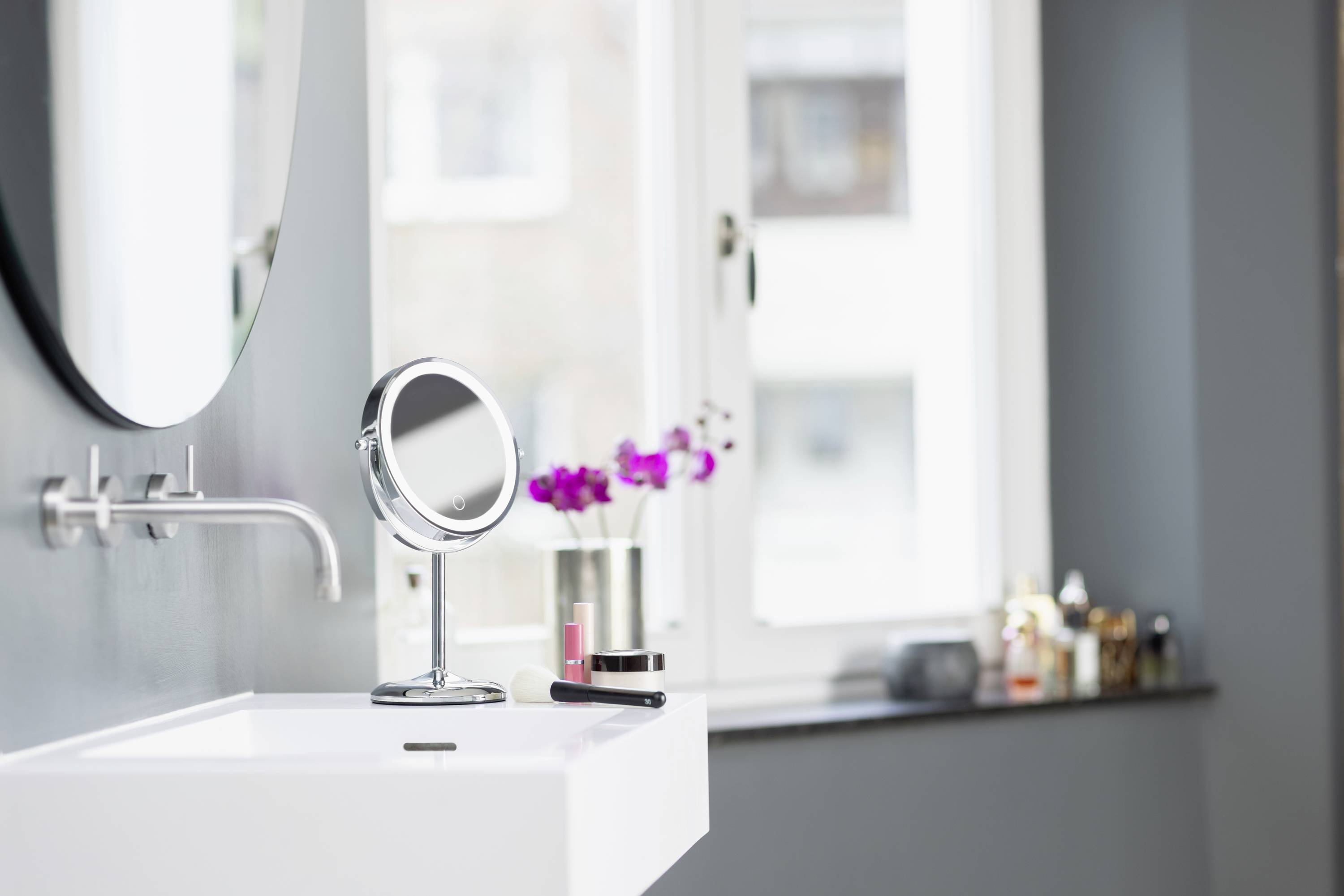 Ein stilvolles Badezimmer mit einem runden Spiegel über einem weißen Waschbecken. Auf dem Beckenrand steht ein Vergrößerungsspiegel.