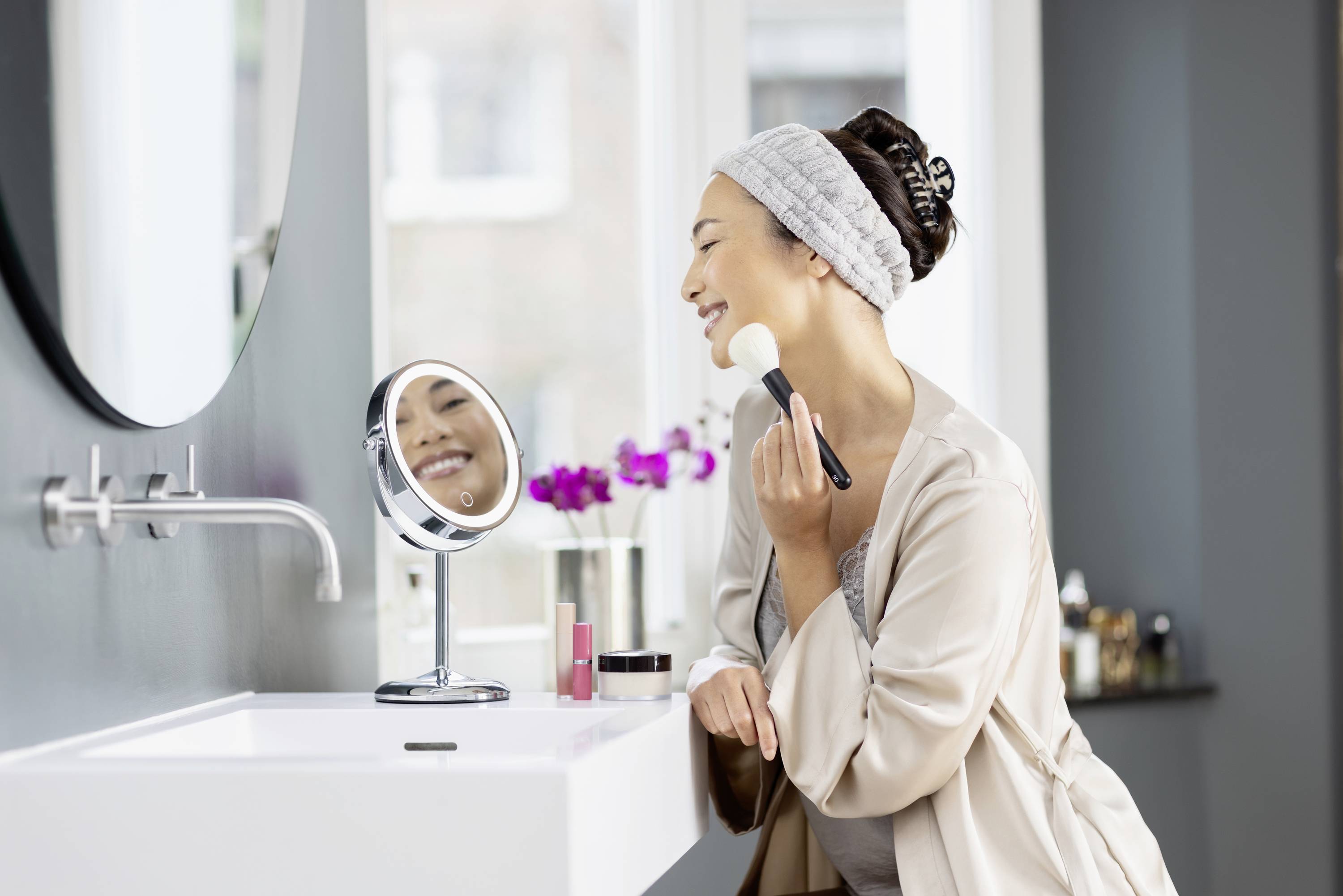 Eine Frau trägt lächelnd Make-up auf ihr Gesicht auf, während sie in einem Badezimmer vor einem Spiegel steht.