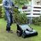 Eine Person mäht mit einem elektrischen Rasenmäher auf einer grünen Wiese in einem Garten. Im Hintergrund sind Büsche und ein Haus zu sehen.