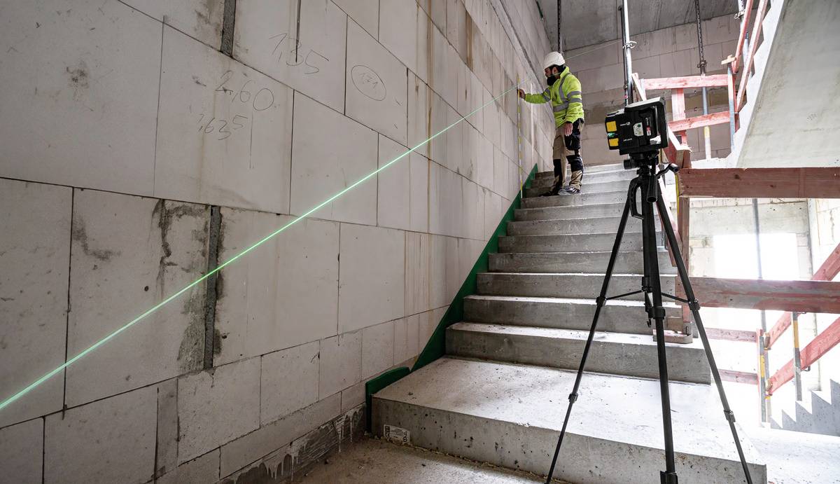Ein Bauarbeiter auf einer Treppe vermisst die Wand mit einem Laser, der von einem Gerät auf einem Stativ ausgeht.