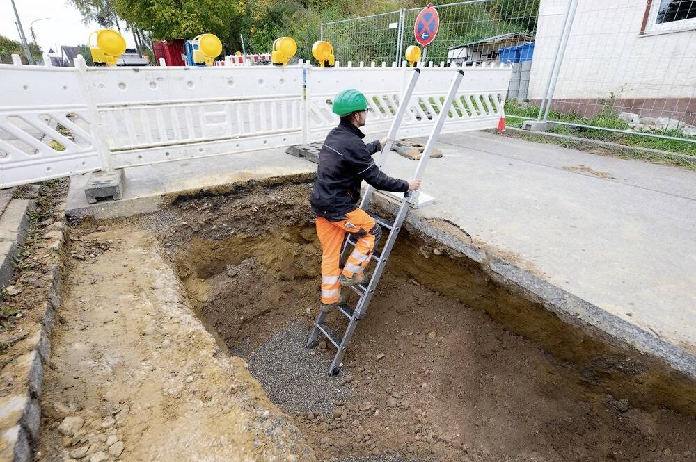 Ein Bauarbeiter mit grünem Helm steigt eine Leiter in eine große Baugrube hinab, umgeben von Absperrungen an einer Straße.