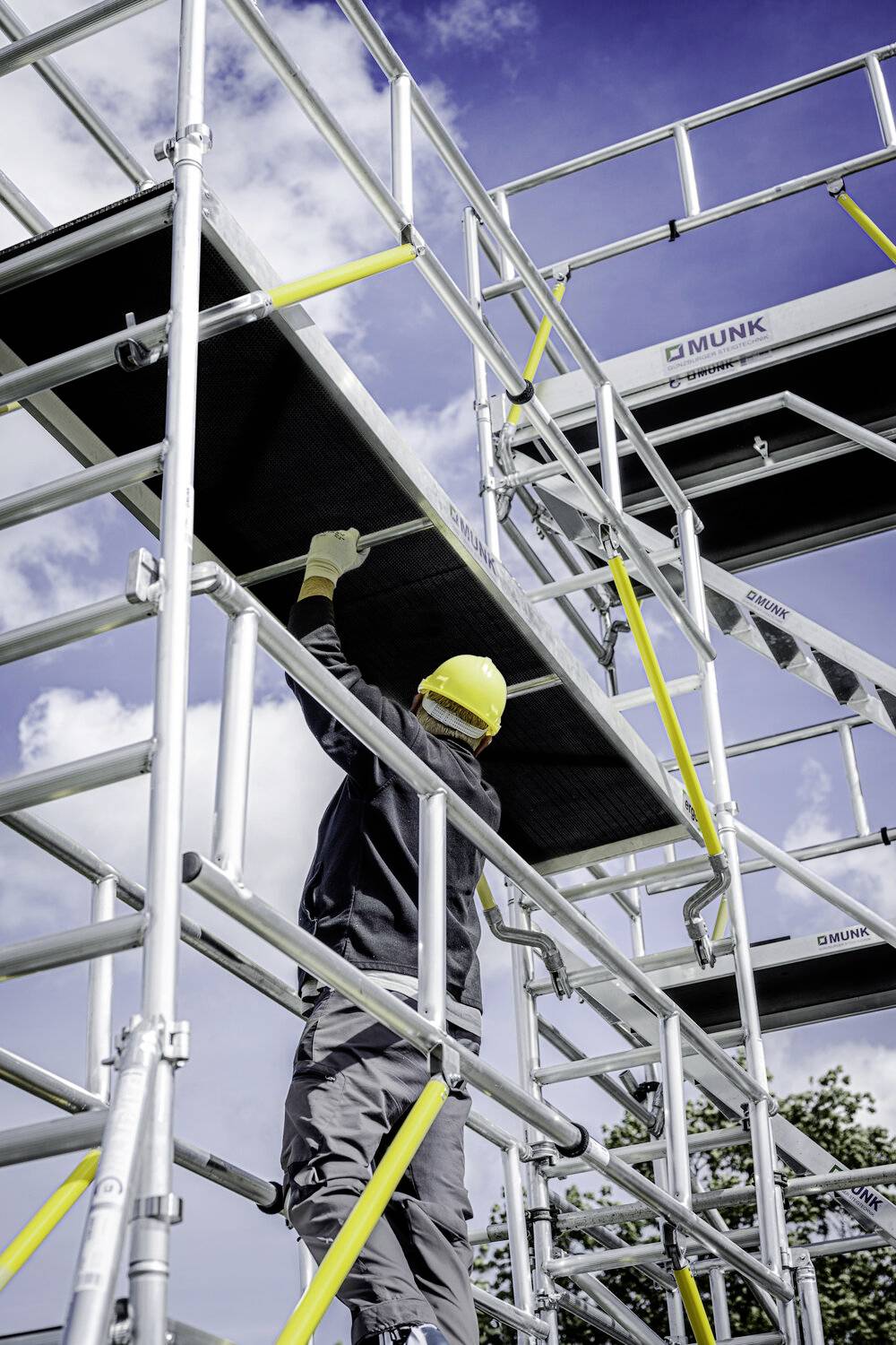 Person arbeitet auf einem hohen Gerüst, trägt Schutzhelm und Handschuhe, befestigt Plattform unter blauem Himmel.