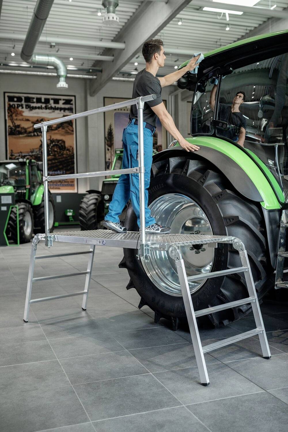 Ein Mann steht auf einer Plattform und reinigt die Frontscheibe eines großen grünen Traktors in einer Ausstellungsraumumgebung.