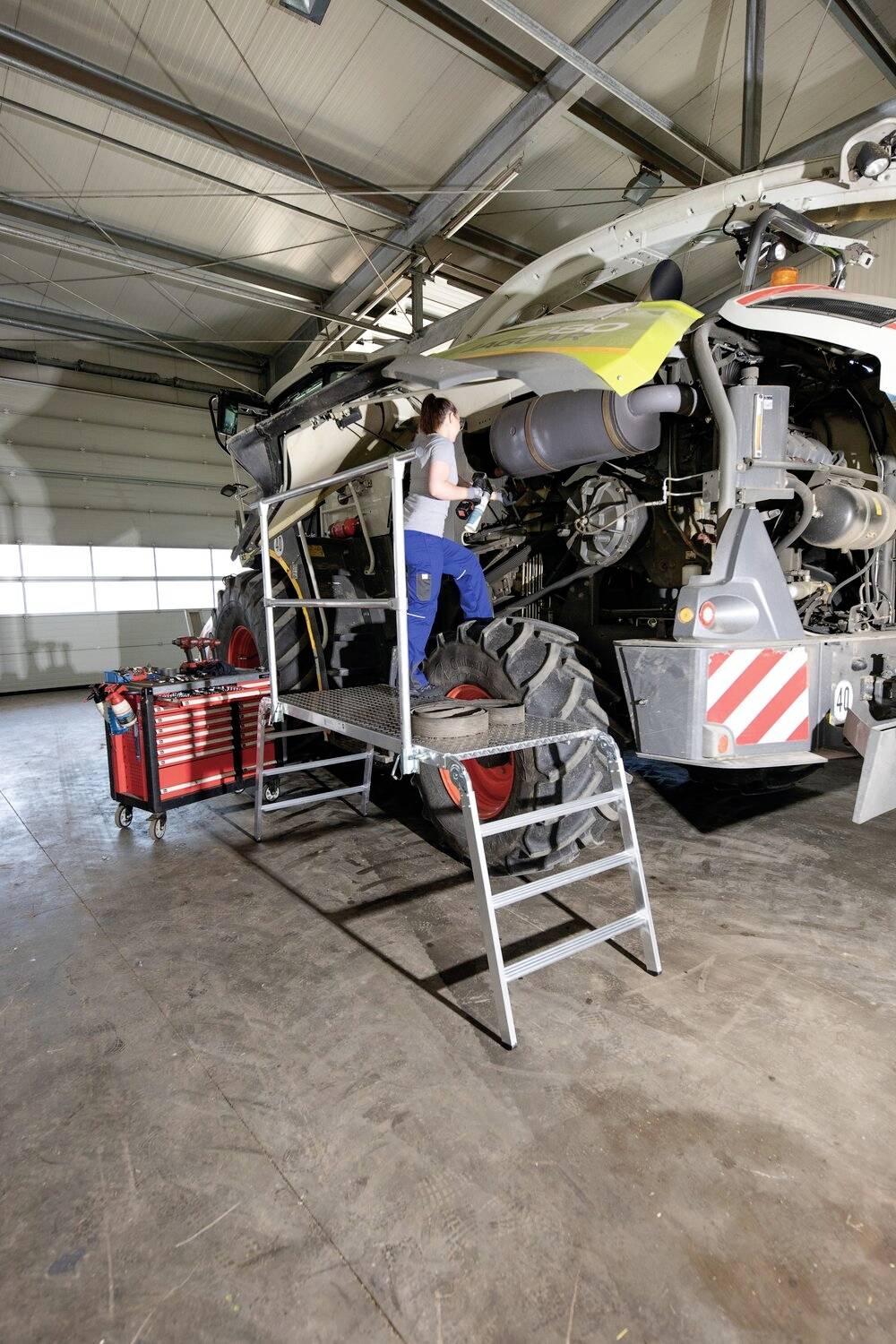 Eine Person arbeitet in einer Werkstatt an einem großen landwirtschaftlichen Fahrzeug, auf einer Leiter stehend, mit Werkzeugen in der Nähe.