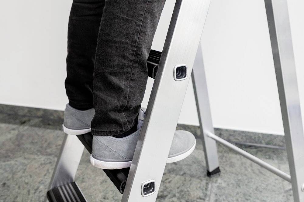 Eine Person steigt eine silberne Leiter hinauf. Man sieht die Beine in schwarzen Hosen und grauen Schuhen. Der Boden ist gefliest.