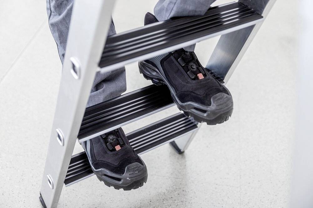 Eine Person trägt schwarze Sicherheitsschuhe und steigt eine silberne Metallleiter hinauf. Die Umgebung ist hell und schlicht.