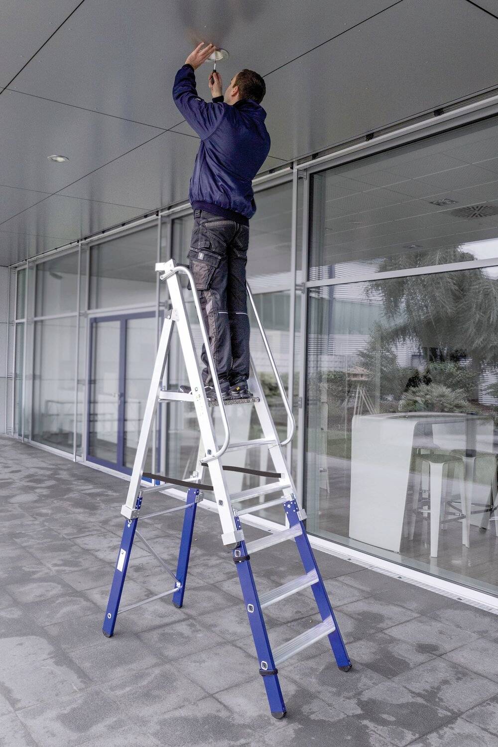 Eine Person steigt auf eine Leiter, um an einer Decke draußen an einem modernen Gebäude zu arbeiten.