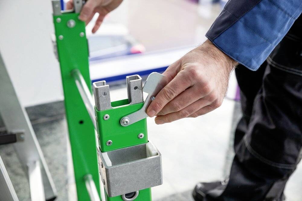 Eine Person justiert den Hebel einer grünen Metallleiter. Die Szene zeigt den Fokus auf den Bewegungsmechanismus für Stabilität.