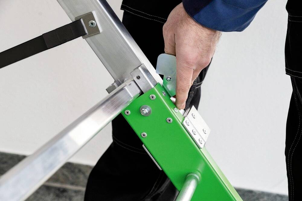 'Hand sichert grüne Trittleiter durch Betätigung einer Klemme.'