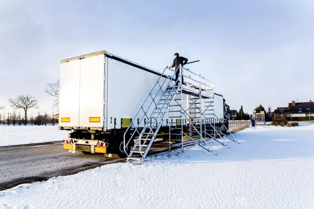 Ein Mann steht auf einer Treppe neben einem großen, weißen LKW-Anhänger in schneebedeckter Landschaft.