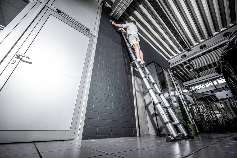 Eine Person steht auf einer hohen Leiter und arbeitet an einer Decke in einer modernen Garage oder Werkstatt mit Metallstrukturen.