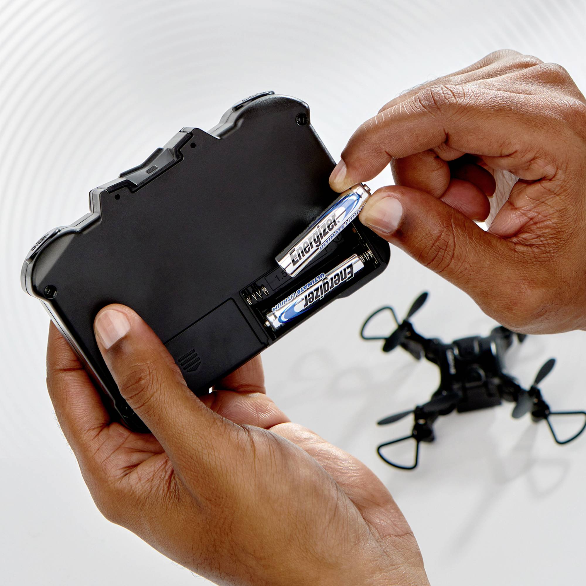 Eine Hand setzt Batterien in die Rückseite einer Fernbedienung ein. Ein kleiner schwarzer ferngesteuerter Hubschrauber ist zu sehen.