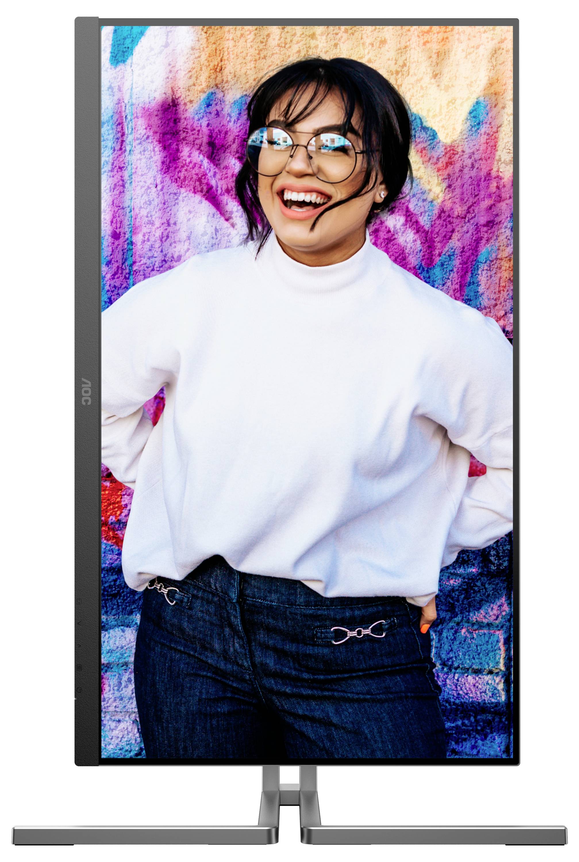 Eine lachende Person steht vor einer bunten Wand. Sie trägt eine Brille, einen weißen Pullover und Jeans.