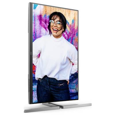 Eine lächelnde Person vor einer bunten, gesprühten Wand. Die Person trägt eine Brille, einen weißen Pullover und dunkle Jeans.