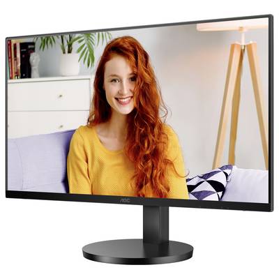 Eine lächelnde Person mit langen roten Haaren sitzt auf einem Sofa vor einem Monitor. Im Hintergrund sind ein Lampenschirm und Pflanzen zu sehen.