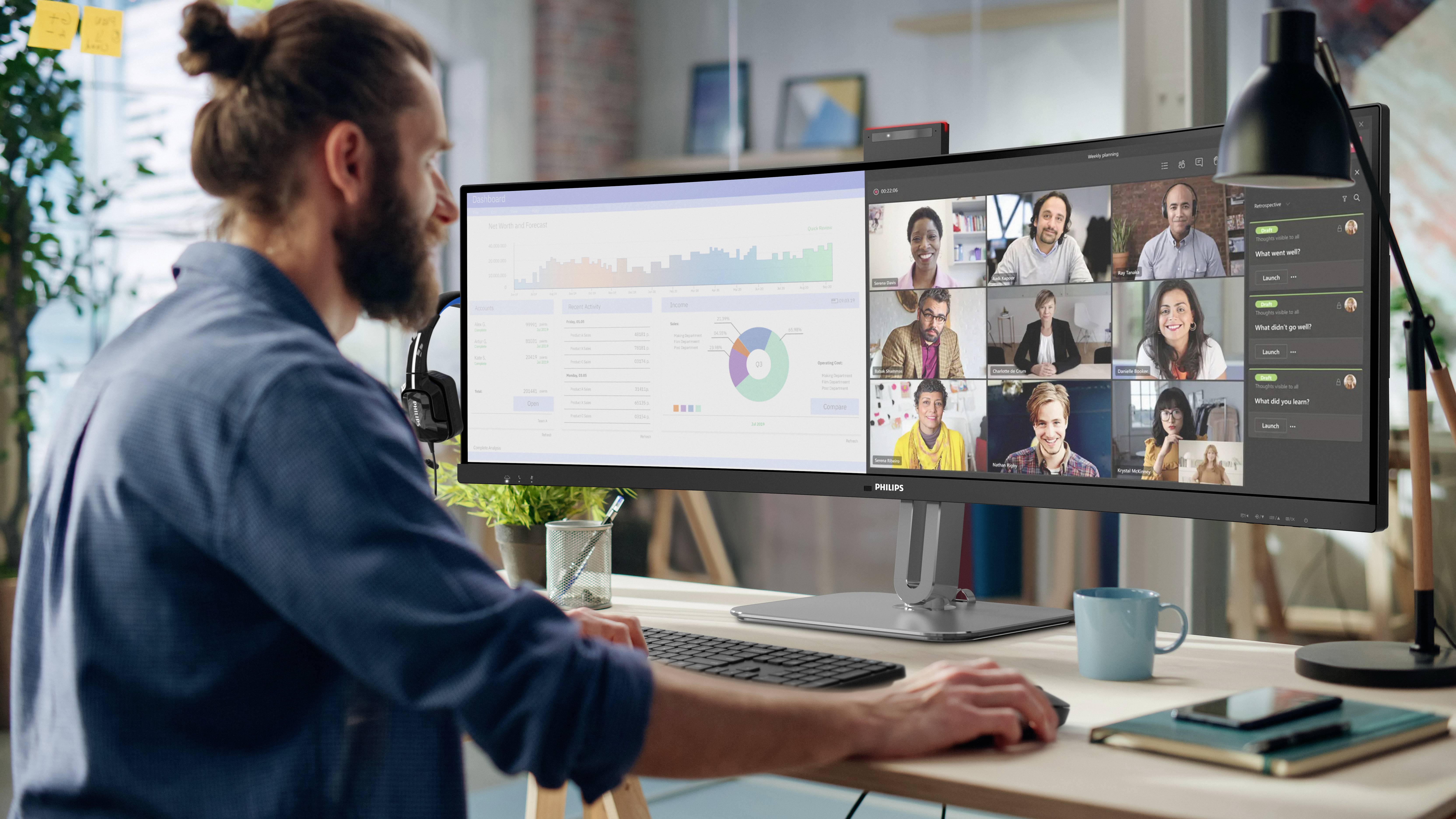 Eine Person sitzt an einem Schreibtisch, arbeitet am Computer mit einem großen Bildschirm. Auf dem Monitor sind Diagramme und ein Videoanruf mit mehreren Personen zu sehen.