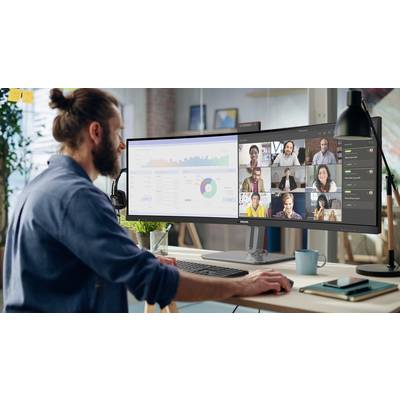 Eine Person sitzt an einem Schreibtisch, arbeitet am Computer mit einem großen Bildschirm. Auf dem Monitor sind Diagramme und ein Videoanruf mit mehreren Personen zu sehen.