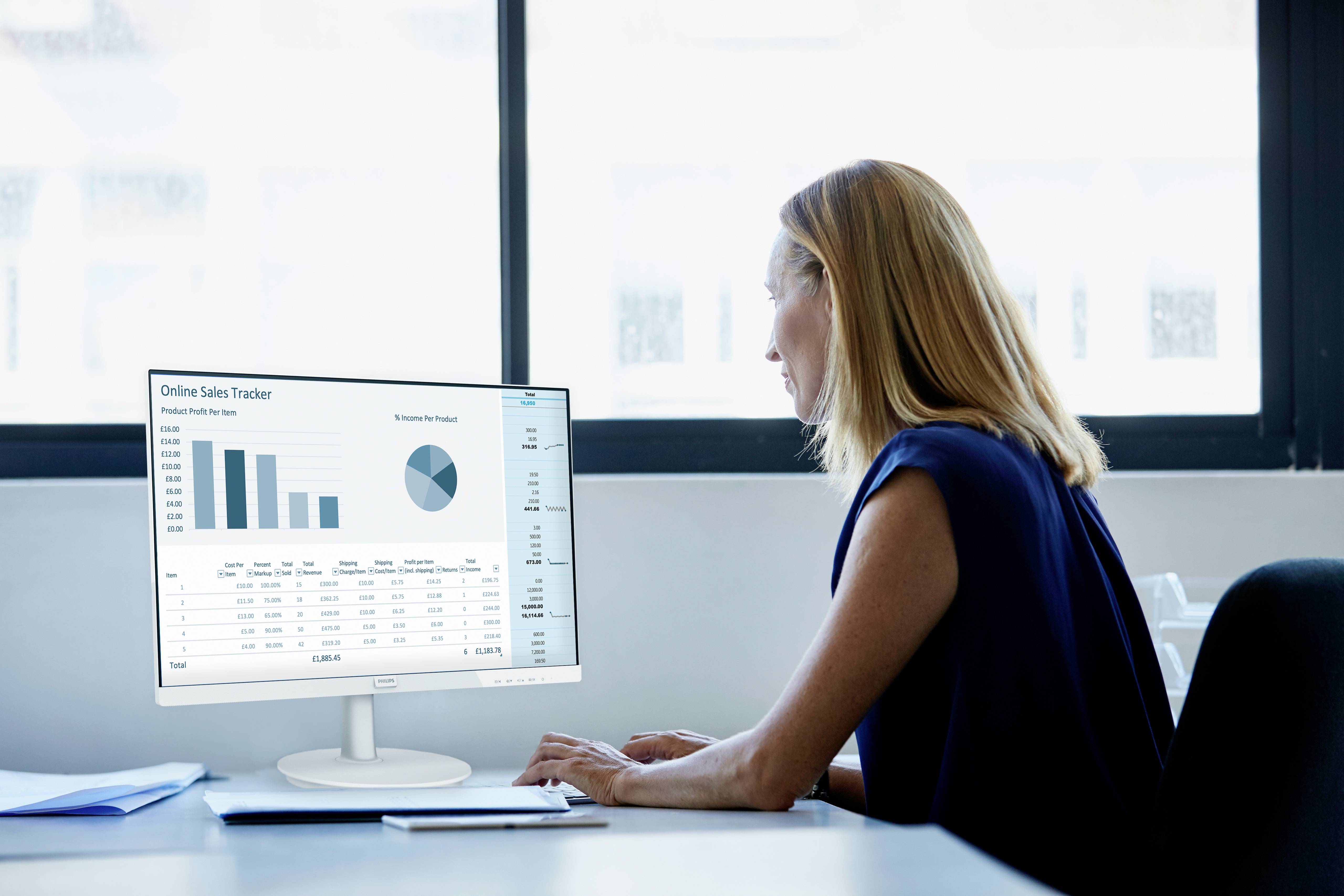 Eine Frau sitzt an einem Schreibtisch, blickt konzentriert auf einen Computermonitor, der Verkaufsdaten-Diagramme und Tabellen zeigt.